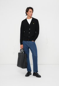 Black knitted cardigan with white buttons, layered over a white shirt. Paired with blue jeans and black shoes, holding a black tote bag.