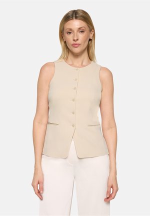 Blonde Frau trägt eine ärmellose beige Weste mit Knöpfen und weiße Hose, steht vor einem schlichten hellen Hintergrund.