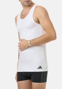 White tank top made of cotton, featuring a round neck and sleeveless design. Black Adidas logo near the hem. Worn with black athletic shorts.