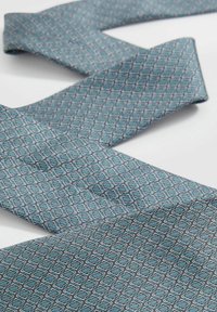 Cravate bleue avec un motif géométrique de losanges entrelacés dans des tons clairs et foncés, fabriquée en tissu lisse, avec des extrémités pointues.