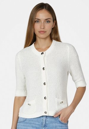 Junge Frau mit langen hellbraunen Haaren, die eine weiße strukturierte Kurzarm-Strickjacke mit silbernen Knöpfen und blaue Jeans trägt.