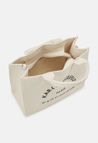 Beige canvas schoudertas met dubbele hengsels, open bovenkant, binnenzak met ritssluiting en bedrukte tekstdetails. Stevige structuur met meerdere compartimenten.