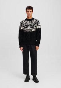 Pull en tricot noir avec un motif géométrique blanc au niveau de l'emmanchure, design en tricot torsadé texturé, associé à un pantalon noir à jambe droite et des chaussures.