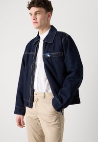 Tumši zila džinsu jaka ar rāvējslēdzēju priekšpusē, kontrastējošām oranžām šuvēm un mazu zilu birku. Valkāta virs baltas krekla un bēšām biksēm.