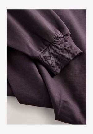 Vijoličasta majica z dolgimi rokavi, opremljena z rebrastimi manšetami in mehkim, teksturiranim materialom. Minimalističen dizajn, brez vidnih logotipov ali vzorcev.