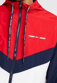 Red, navy, and white windbreaker jacket with zip closure, hood, and logo. Features color-block design, textured fabric, and drawstring details.