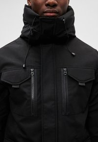 Chaqueta aislante negra con alto cuello, dos bolsillos en el pecho con cremallera y cuerditas exteriores para ajuste. Textura suave y mate.