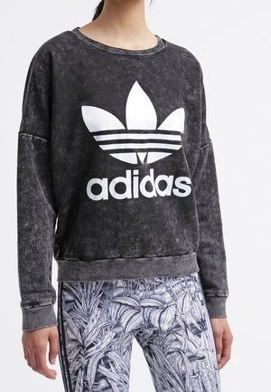 Personne portant un sweat-shirt Adidas noir avec logo blanc et des leggings à motifs feuilles sur fond blanc.