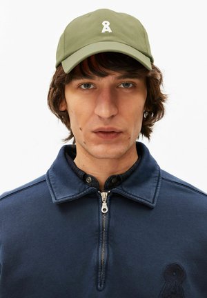 Jonge man met halflang bruin haar, die een olijfgroene beanie en een marineblauwe jas met ritssluiting draagt, tegen een witte achtergrond.