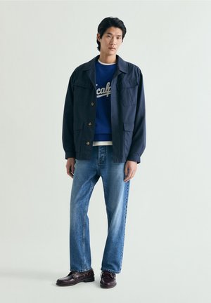 Jeune homme debout portant une veste bleu marine, un sweat-shirt bleu avec texte blanc, un jean bleu clair et des chaussures marron foncé sur fond uni.