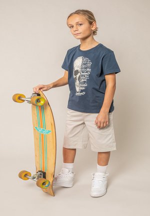 Jonge jongen met een marineblauw T-shirt met schedelprint en beige korte broek, die een houten longboard-skateboard met gele wielen vasthoudt, staand tegen een lichte achtergrond.