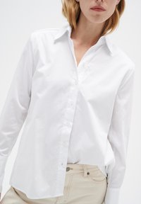 Camisa blanca de botones con cuello, hecha de una suave tela de algodón. Presenta mangas largas y un corte relajado. Combinada con pantalones beiges.