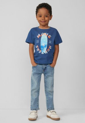 Niebieski bawełniany t-shirt z grafiką deski surfingowej, zestawiony z jasnoniebieskimi dżinsami i białymi trampkami z brązowymi akcentami na podeszwie.