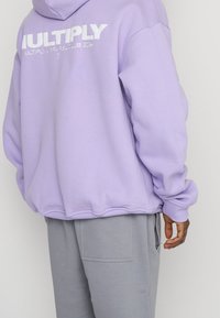 Felpa lavender con vestibilità ampia, caratterizzata da un grande logo bianco "MULTIPLY" sul retro e polsini a coste, abbinata a pantaloni tuta grigi.