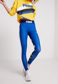Modré legíny s logem PUMA, v kombinaci se žlutou mikinou s geometrickými pruhy v černé a modré. Bílé tenisky dotvářejí vzhled.