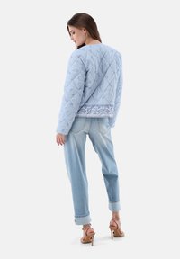 Hellblaue gesteppte Jacke mit Paisleymuster-Akzenten, getragen über hellblauen hochgekrempelten Jeans und hautfarbenen Absatzsandalen, von hinten gesehen.