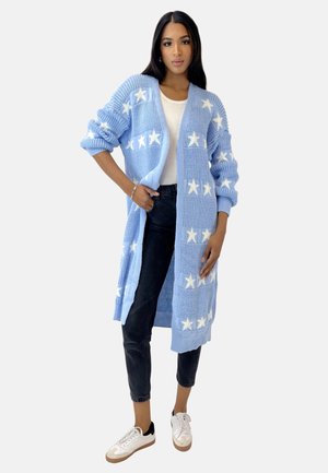 Cardigan lavorato a maglia di colore blu chiaro con motivi a stelle bianchi; aperto davanti, maniche lunghe, tessuto strutturato. Indossato sopra una maglietta bianca e jeans neri.