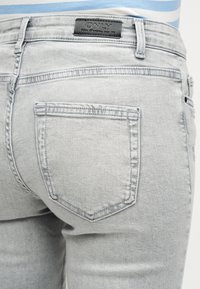 Ανοιχτό γκρι τζιν με πίσω τσέπη και μαύρη ετικέτα μάρκας που γράφει "ONLY", φορεμένα από ένα άτομο με μπλε και λευκή ριγέ πουκάμισο.