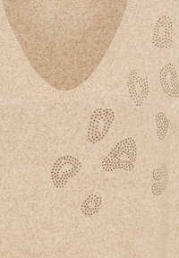 Beige V-Ausschnitt-Pullover mit kleinen metallischen Nieten, die abstrahierte Leopardenflecken auf dem Stoff bilden.