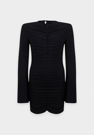 Femme portant une robe mini noire ajustée à fronces, manches longues et bottines noires, debout devant un fond clair uni.