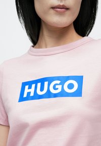 Lyserød T-shirt med rund halsudskæring og en blå rektangulær logo, der lyder "HUGO" i hvide, fede bogstaver. Blød stoftekstur.