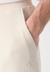 Pantaloni beige con texture a nido d'ape, dotati di una tasca laterale e un logo ricamato discreto. Tessuto morbido e cintura a coste.