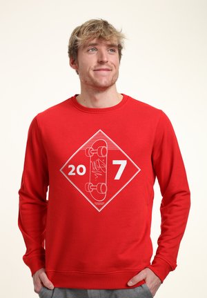 Felpa rossa con maniche lunghe, caratterizzata da un motivo grafico bianco di uno skateboard e dai numeri "20" e "7" all'interno di una forma a diamante.