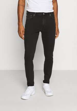 Jeans neri skinny in denim, con un design a cinque tasche, chiusura con zip e bottone. Indossati con sneakers bianche e una camicia bianca.