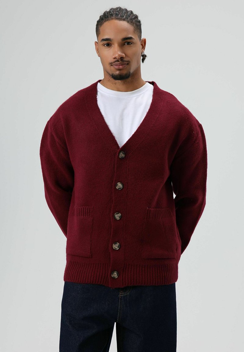 Cardigan bordeaux, texture en maille, encolure en V, six boutons marron, poignets côtelés et deux poches avant. Porté sur un t-shirt blanc et un jean foncé.