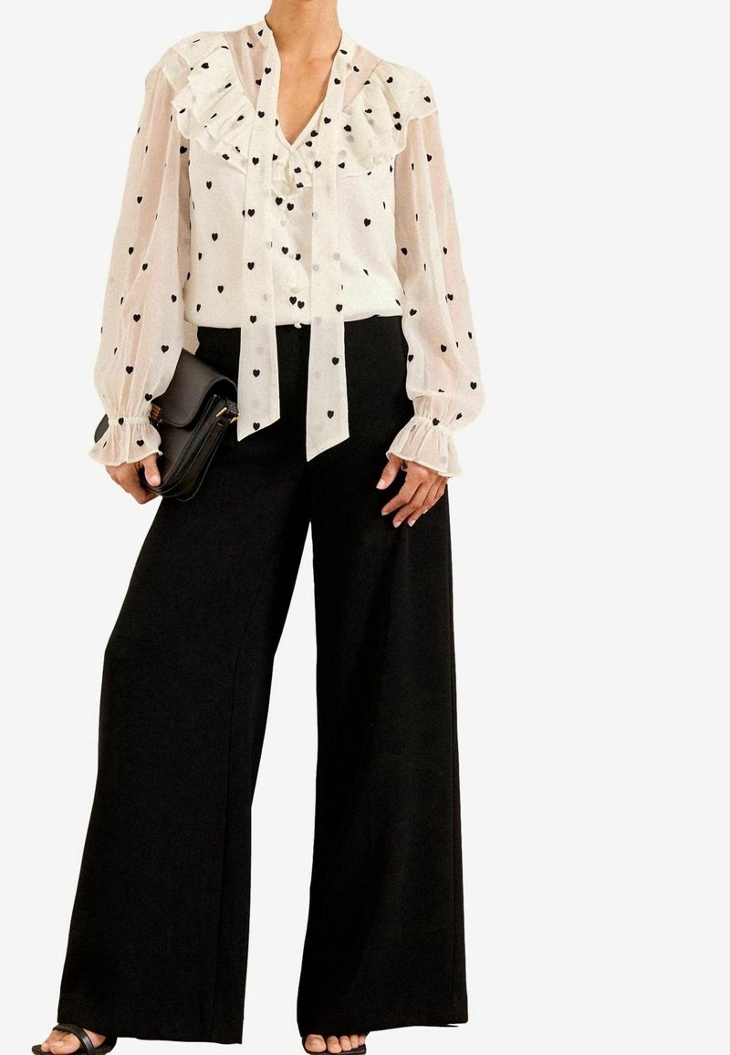 Elle porte un chemisier blanc transparent avec des motifs de cœurs noirs, un col à volants, des manches longues, un pantalon large noir, une pochette noire et des sandales noires.