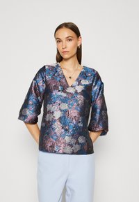 Top floral violet et bleu avec un décolleté en V, des manches trois-quarts, en tissu texturé brillant, et une coupe décontractée. Assorti à un pantalon bleu clair.