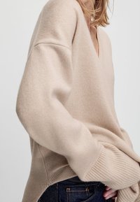 Vrouw in een beige oversized V-halstrui met geribbelde manchetten, gecombineerd met donkerblauwe denim jeans, gezien vanaf de zijkant.