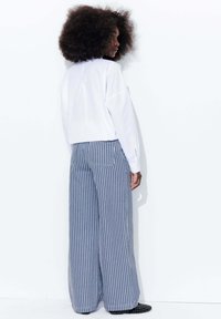 Chemise blanche à manches longues, coupée au-dessus de la taille, associée à un pantalon large à rayures bleues et blanches, présentant deux poches arrière et un tissu texturé.