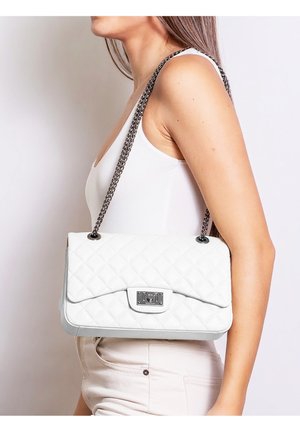 Donna con top bianco senza maniche e pantaloni chiari che porta una borsa a tracolla bianca trapuntata con catena argento e chiusura metallica.
