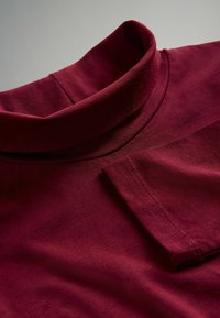 Maglione a maniche lunghe bordeaux con collo a dolcevita, posato piatto su una superficie, con dettagli del collo e delle maniche piegate ben visibili.