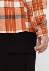 Camisa de franela a cuadros en naranja, blanco y gris, con puños abotonados y corte relajado. Se ven pantalones cargo negros con un bolsillo lateral.
