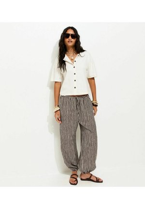 Woman wearing white buttoned shirt, black and white vertical striped loose pants, brown sandals, sunglasses, and multiple bracelets stands against white wall.