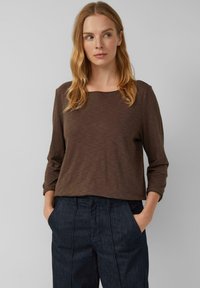 Kobieta z blond włosami do ramion, ubrana w brązową bluzkę z długim rękawem i ciemne dżinsy, stojąca z rękami w kieszeniach, patrząca w bok.