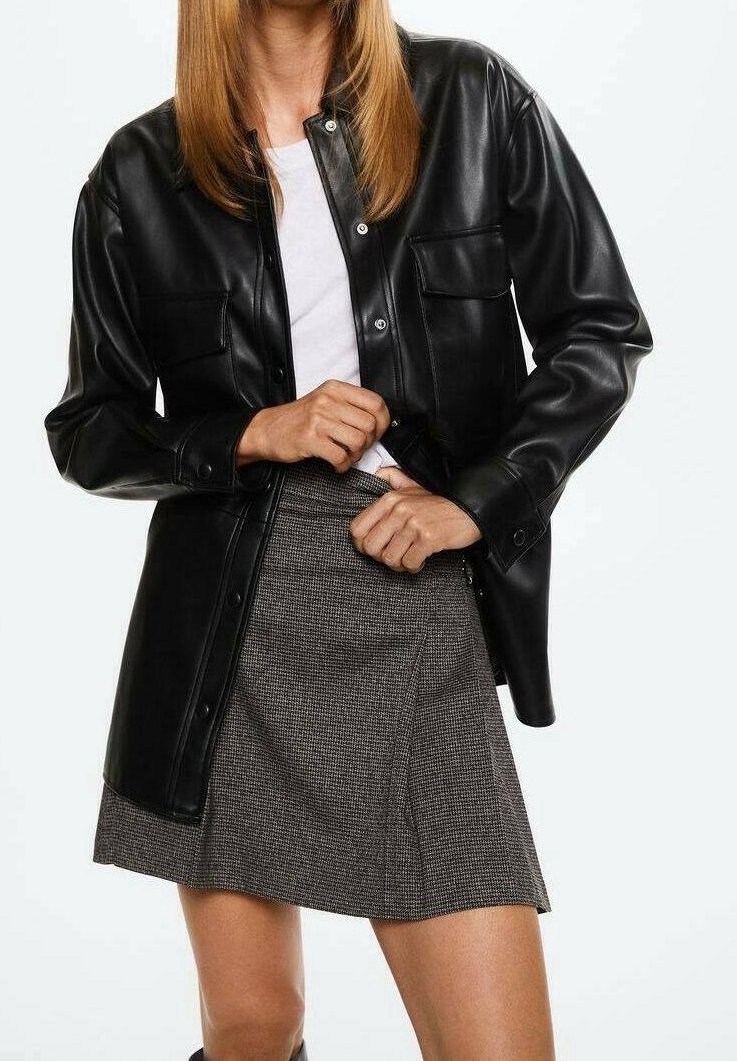 Femme portant une veste en cuir noire boutonnée sur une chemise blanche et une jupe grise à carreaux, ajustant la veste à la taille.