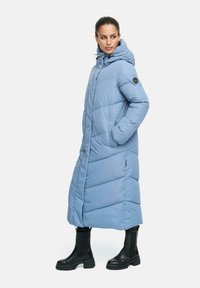 Cappotto lungo imbottito di colore blu chiaro con cappuccio, design trapuntato, tasche laterali e un logo circolare nero sulla manica, indossato con stivali neri alla caviglia.