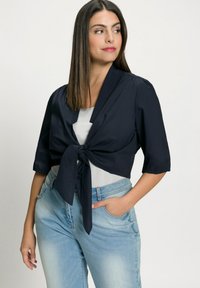 Navy cropped tie-front jacket with short sleeves, made from lightweight fabric. Paired with light blue denim, featuring a pocket on the side.