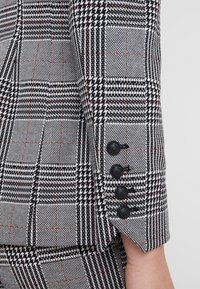 Manche et pantalon d'un costume à motif pied-de-poule noir et blanc avec quatre boutons noirs sur le poignet de la manche, porté sur le bras d'une personne.