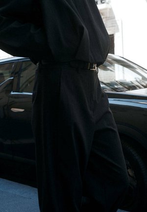 Personne vêtue d’un pantalon noir ample et d’une chemise noire, debout à côté d’une voiture sombre dans une rue de la ville.