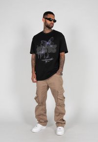 Svart oversized t-skjorte med lilla sommerfuglgrafikk og stilisert tekst, parret med beige cargo-bukser og hvite joggesko. Stilige solbriller er båret.