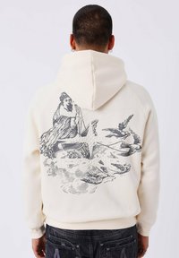 Persoon in een crèmekleurige hoodie met een zwart vintage illustratie van een vrouw, vogels, wolken en decoratieve elementen op de rug.