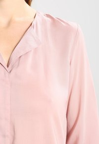 Hellrosa Bluse mit V-Ausschnitt, weicher Textur und lockerer Passform. Verfügt über feine Nähte und minimale Designdetails.