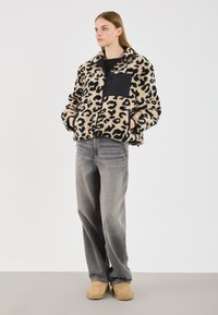 Mladá žena v béžovo-čiernom leopardom fleecovom kabáte, šedých širokých nohaviciach a béžových nazúvacích topánkach, stojí s rukami v vreckách.