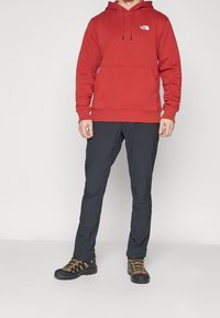 Roter Pullover-Hoodie mit Fronttasche und verstellbarem Kordelzug, kombiniert mit schwarzen Hosen und schwarzen Wanderschuhen mit orangefarbenen Akzenten.