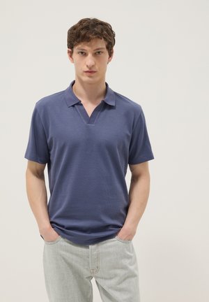 Jonge man met kort bruin haar, die een blauw overhemd met korte mouwen en kraag draagt en lichtgrijze broek, staand met handen in de zakken.