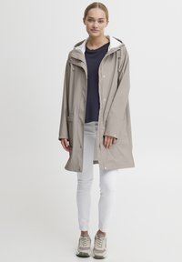 Beige vanntett jakke med hette, med hvit innerforing, trykknapper og to sidelommer, brukt over en marineblå skjorte og hvite bukser.